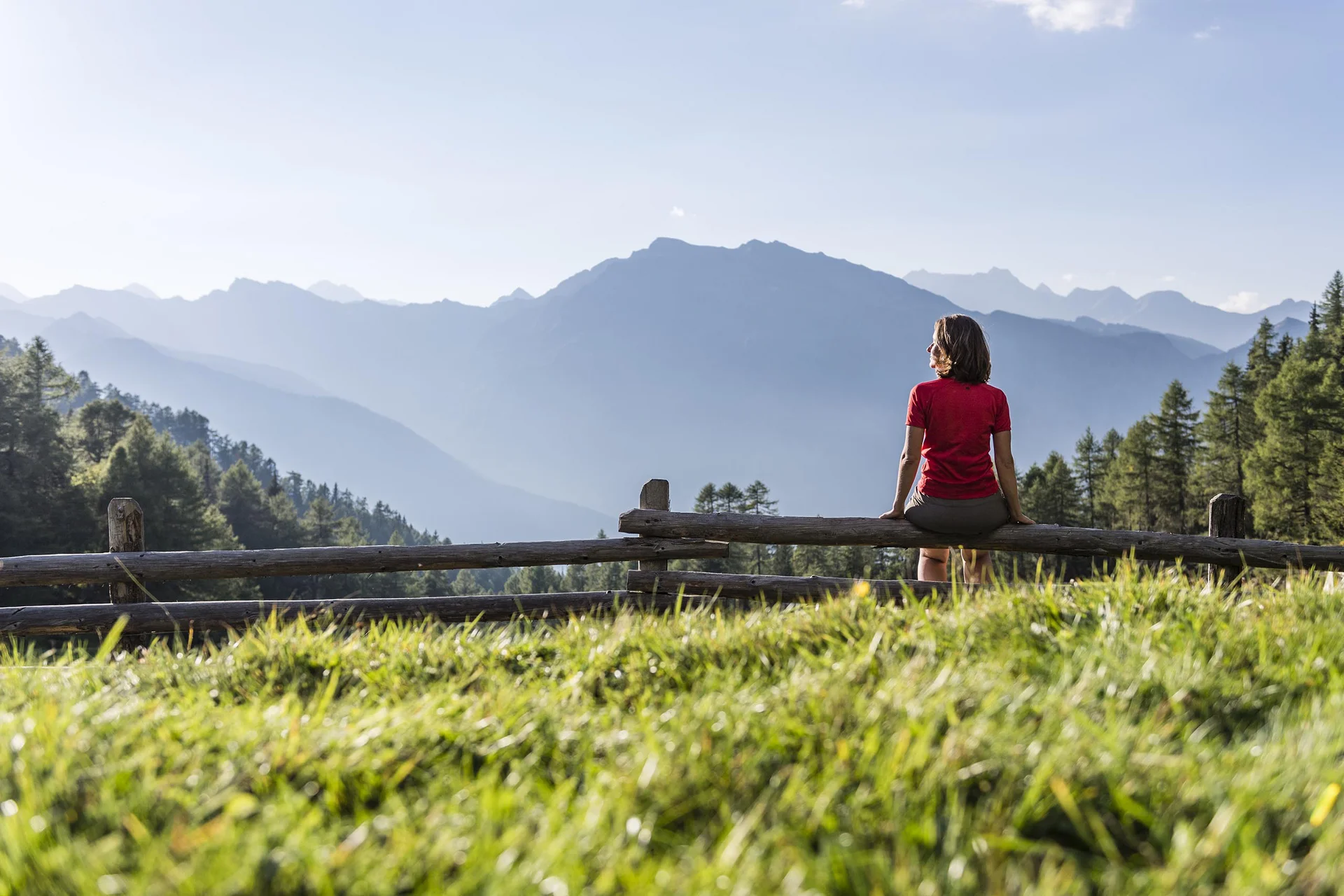 Frau sitzt auf Holzgeländer mit Bergsicht bei sonnigem Wetter