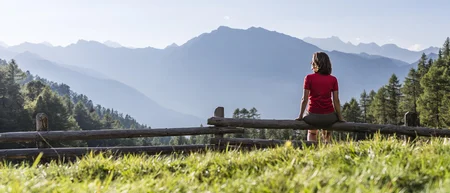 Frau sitzt auf Holzgeländer mit Bergsicht bei sonnigem Wetter