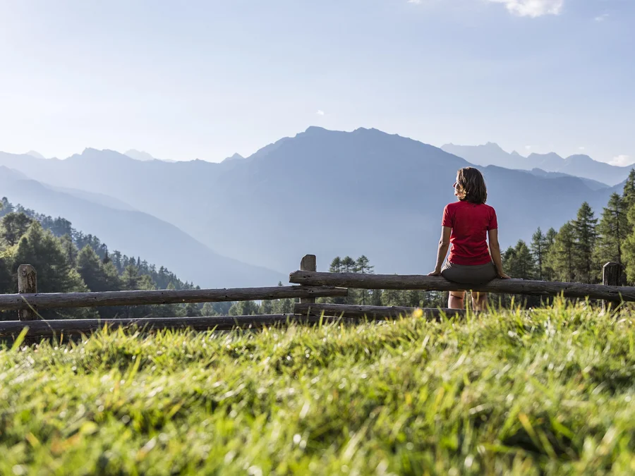 Frau sitzt auf Holzgeländer mit Bergsicht bei sonnigem Wetter